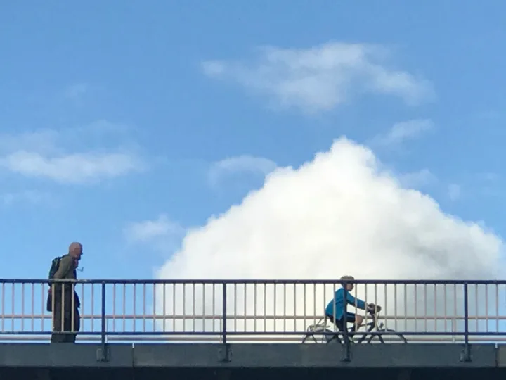 A pedestrian and a cyclist crossing a bridge beneath white clouds.